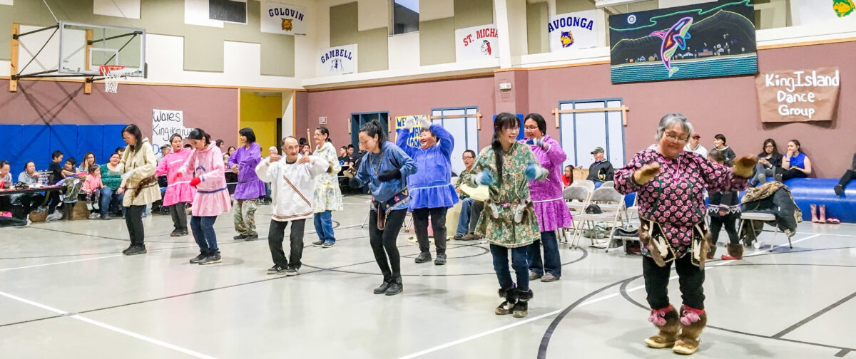 An Alaska Native dance ensemble performs inside a school gymnasium.