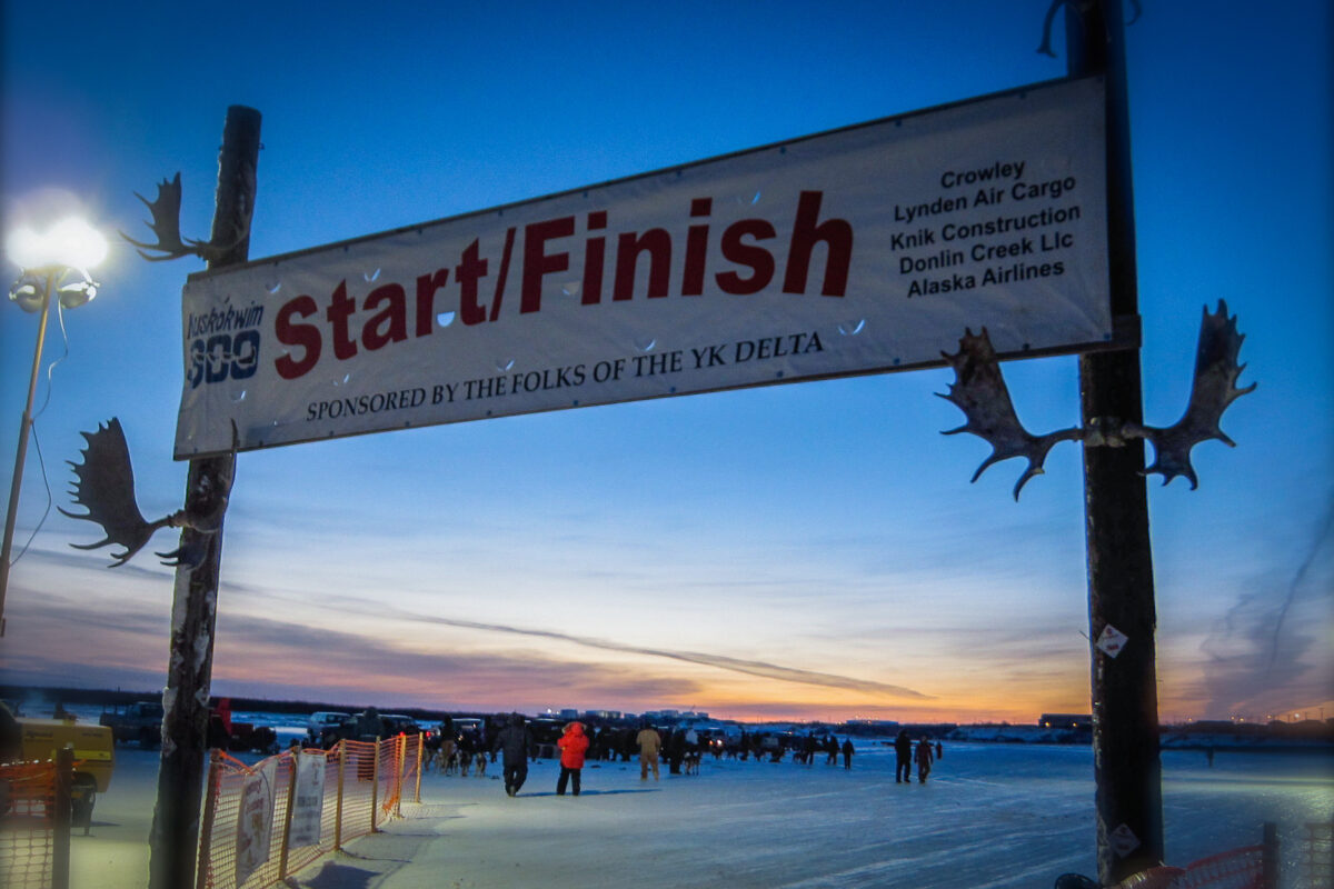 A banner reading "Start/Finish" and adorned with antlers hangs over an icy, snowy field against a colorful sunset.