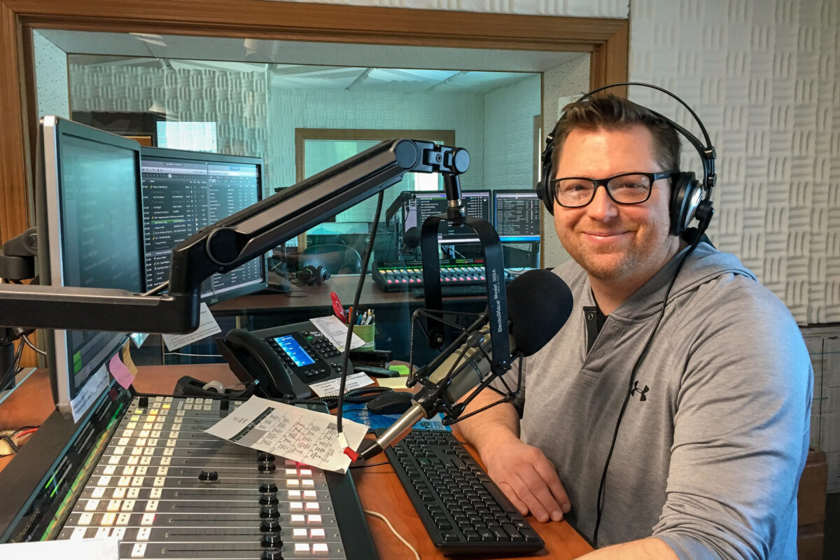 Deejay Michael Burnett, wearing headphones, standing at the microphone and radio sound board in KNOM's main broadcasting studio.