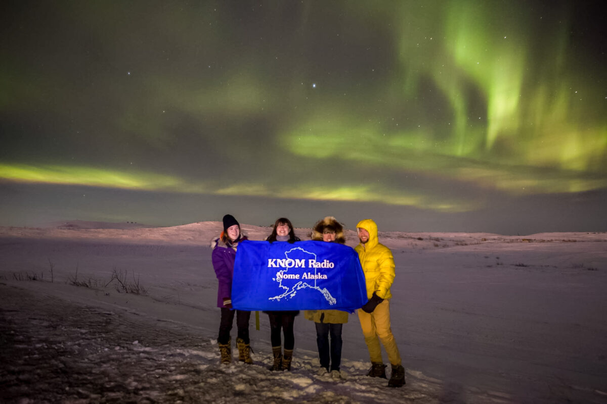 2016-2017 volunteers Karen Trop, Lauren Frost, Tyler Stup, and Davis Hovey hold a KNOM Radio banner under a particularly vivid, green-colored instance of the Northern lights near Nome.