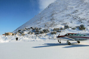 A landscape of Diomede, Alaska, in wintertime: a small airplane sits on a runway made of ice, with village houses nestled at the base of a steep hill in the background.
