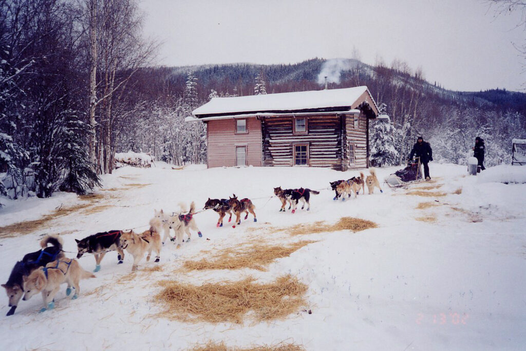A dogsled team departs a log cabin at Slaven's Roadhouse. Photo Credit: National Park Service (2013)