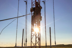 A silhouette of a crewman ascending a radio tower with the sun and tundra landscape behind him.