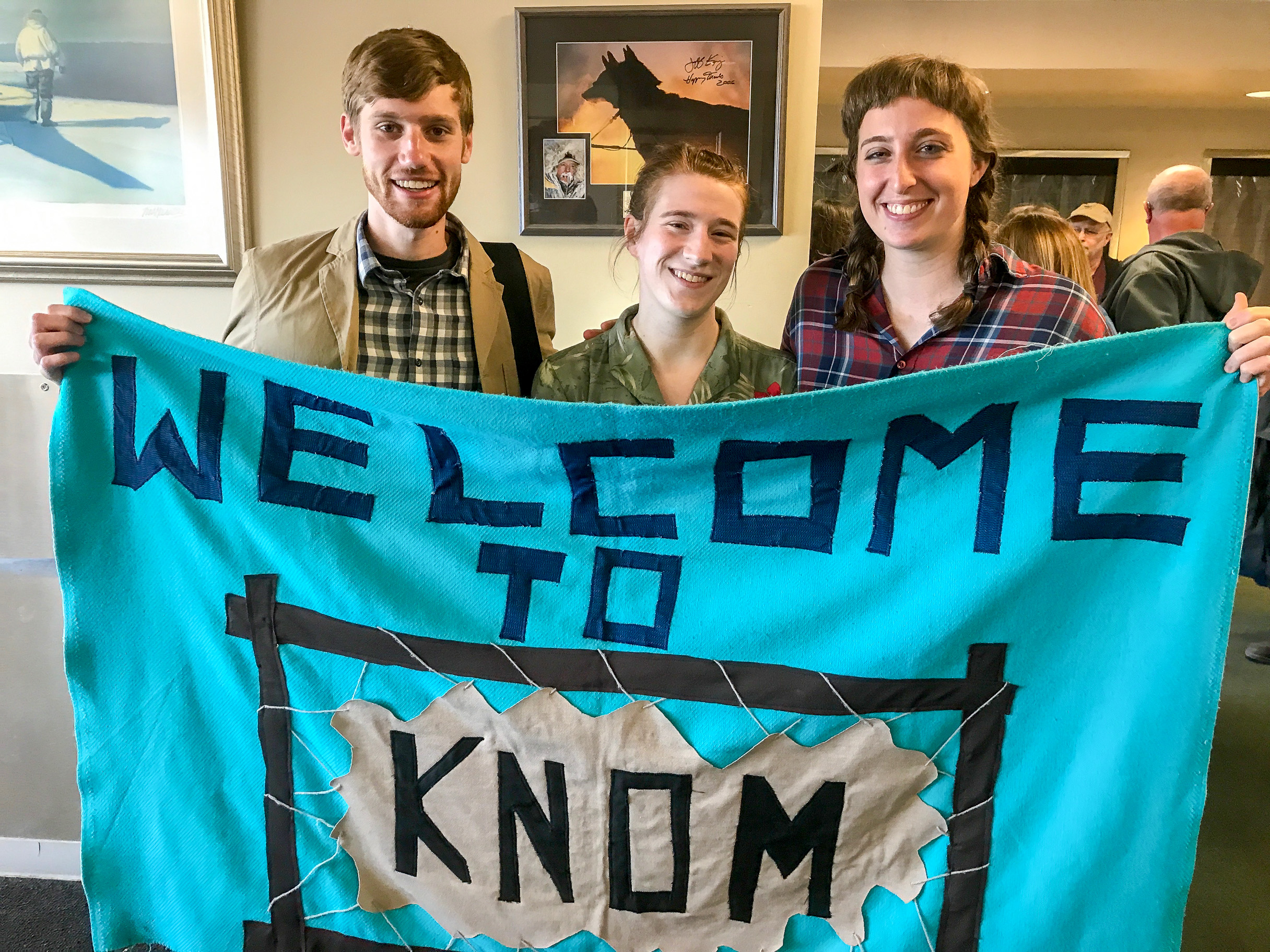 At the Nome airport: Gabe Colombo, Karen Trop, and Zoe Grueskin holding the “Welcome to KNOM” banner that greets newly-arriving volunteers.