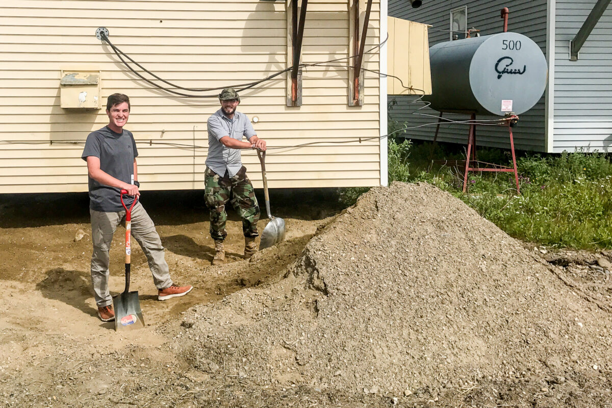 Two men with shovels stand aside a mound of dirt behind KNOM's garage