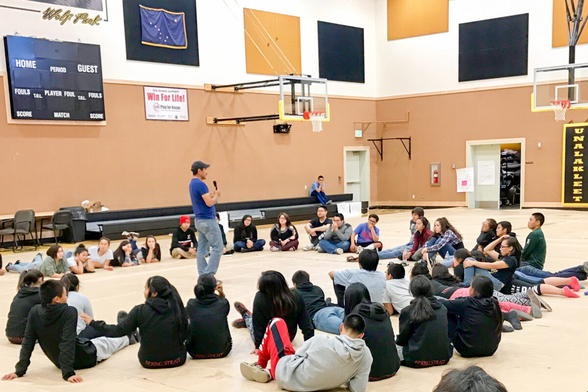 Teenagers sit in a large circle on the floor of a school gymnasium to listen to a presenter.