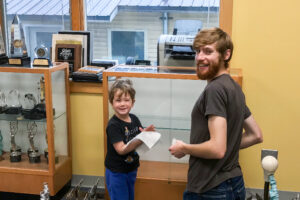 Young child and bearded man, both smiling, help dust display case in lobby of radio station