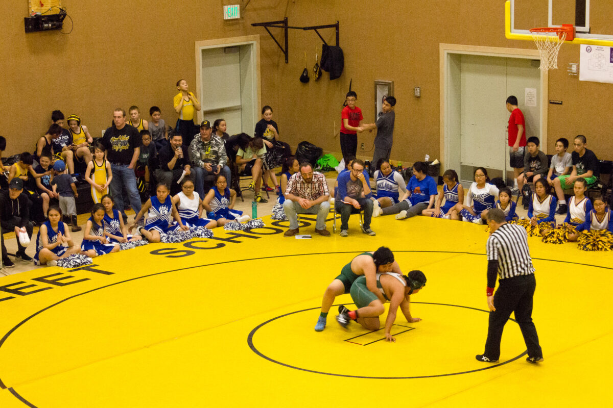 Wrestlers wrestle on a yellow mat surrounded by watching fans