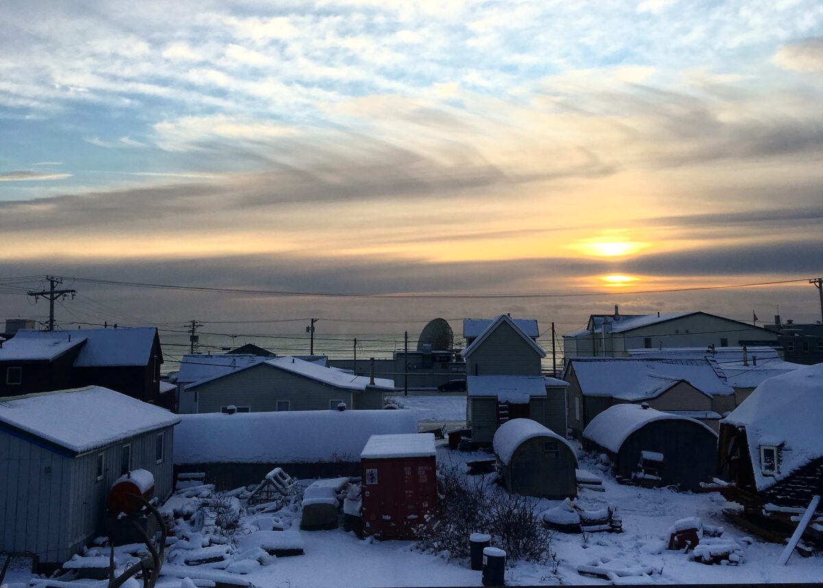 The sun sets over the Bering Sea in the late afternoon. Clouds fill the sky, and snow covers the ground and buildings in Nome