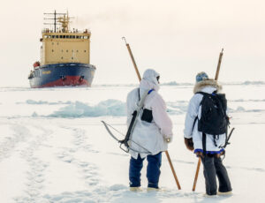 Two people, wearing heavy parkas, hold long poles with iron hooks at the end, standing on sea ice, with a large vessel in the background.