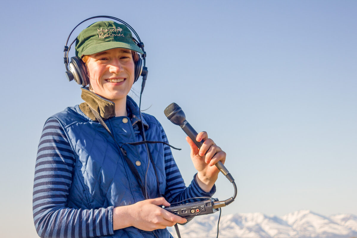 Volunteer Karen Trop, in a blue long-sleeve t-shirt and vest, holds a microphone and small audio recorder, smiling at the camera.