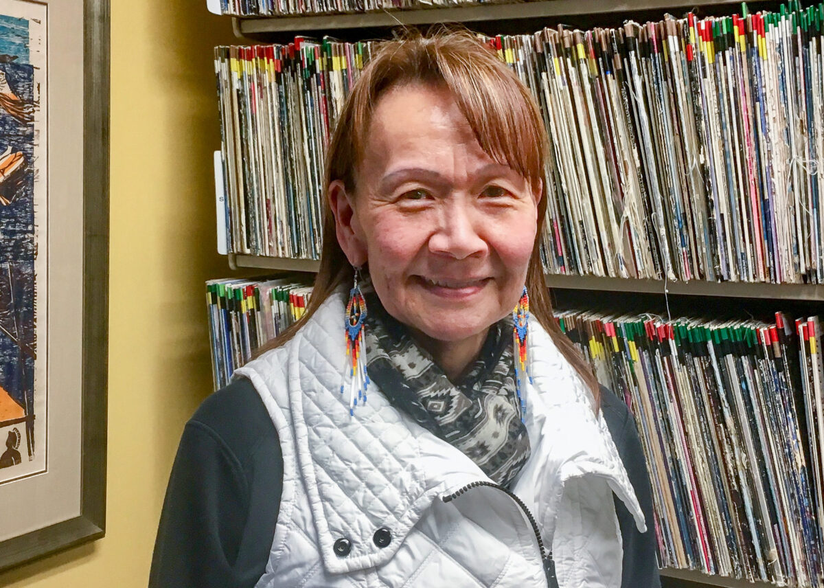Woman in white vest stands next to Alaska Native painting and shelves of vinyl records.