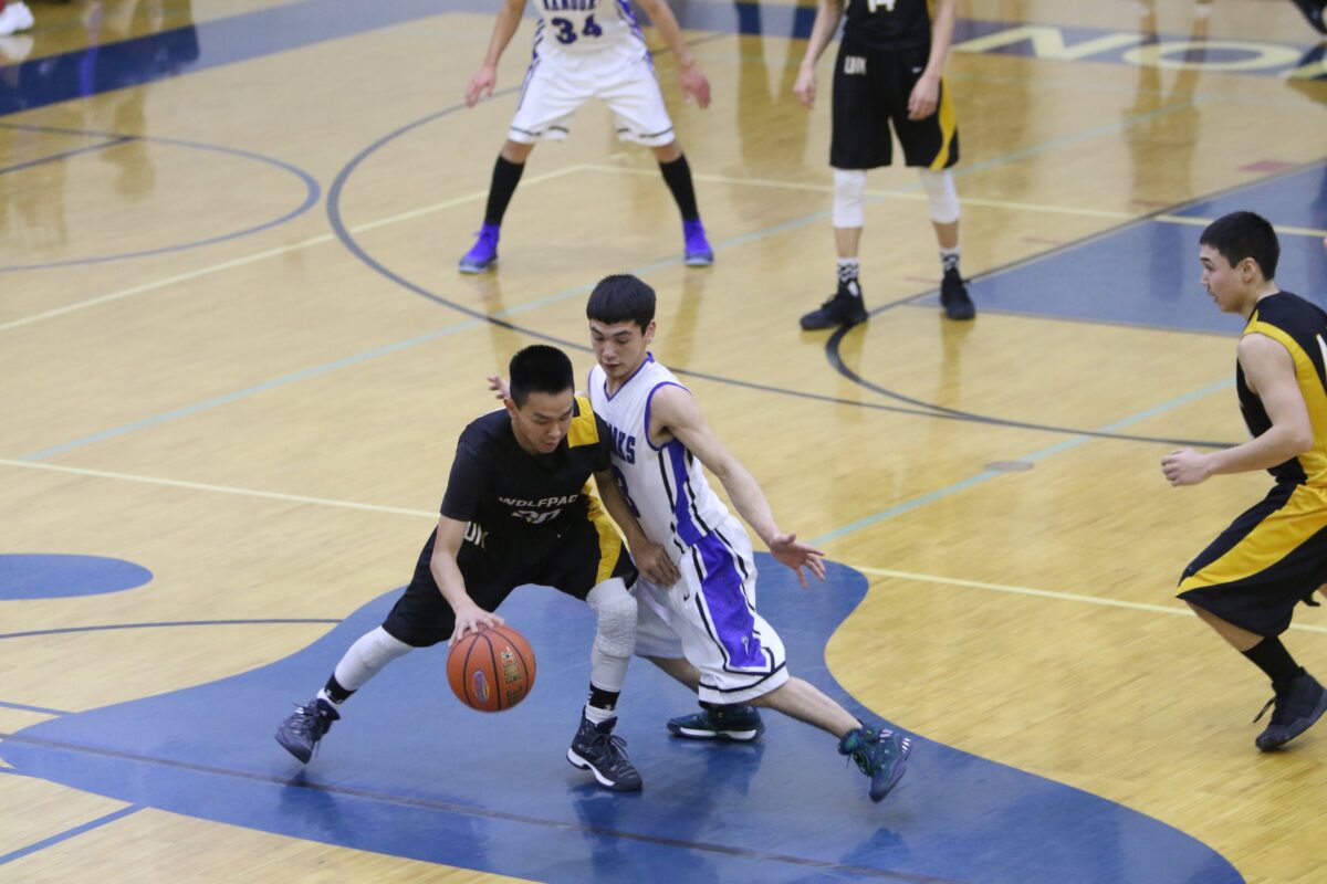 A Unalakleet boy, dressed in black and yellow, dribbles the ball and is blocked by a Nome boy, dressed in white and purple