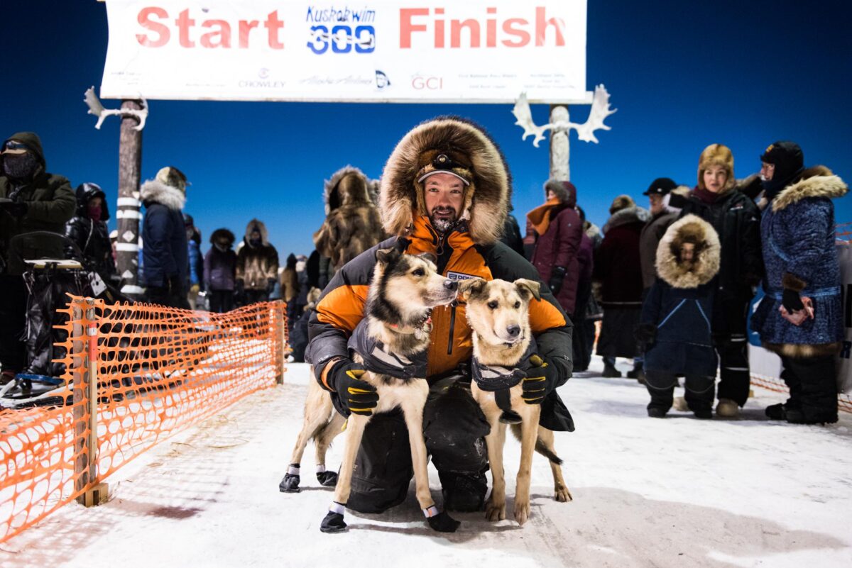 Musher in heavy parka with fur ruff hugs sled dogs under finish line of the Kuskokwim 300 in Bethel, Alaska.