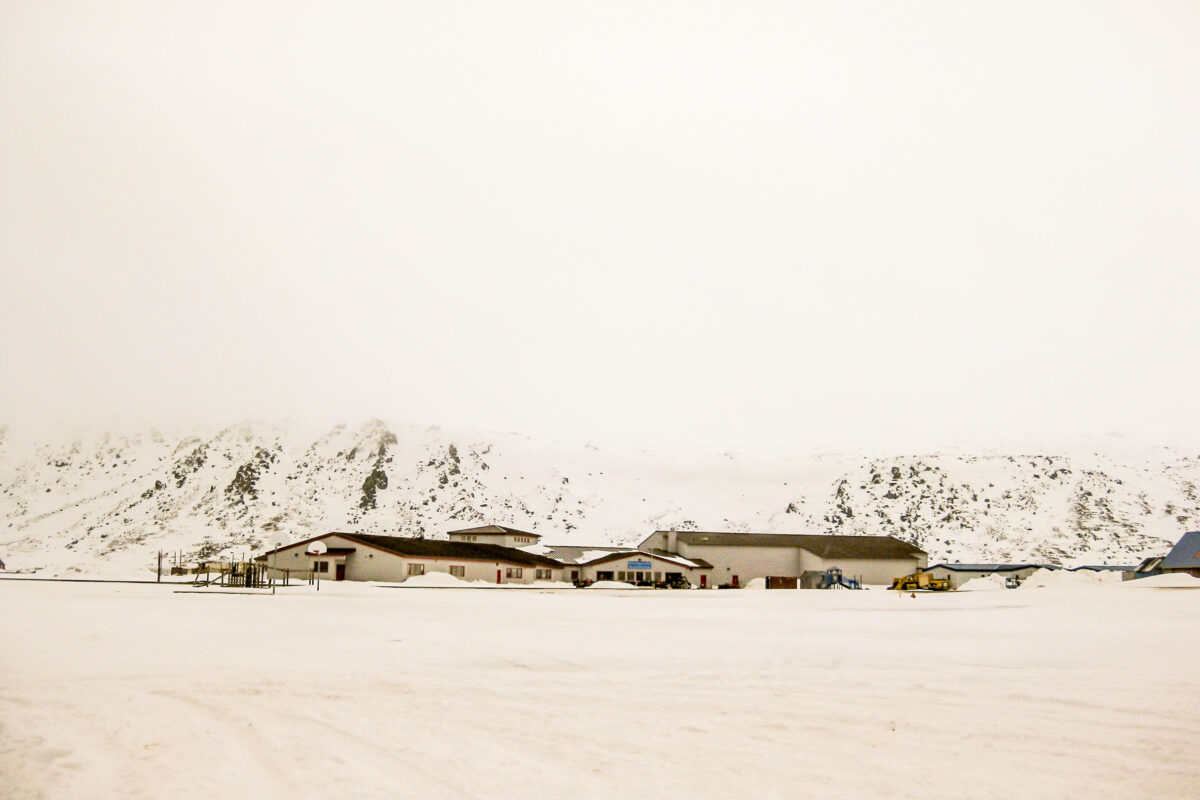 A snowy landscape, distant snowy mountains, low-hanging fog, and a modern school complex in the middle-ground.