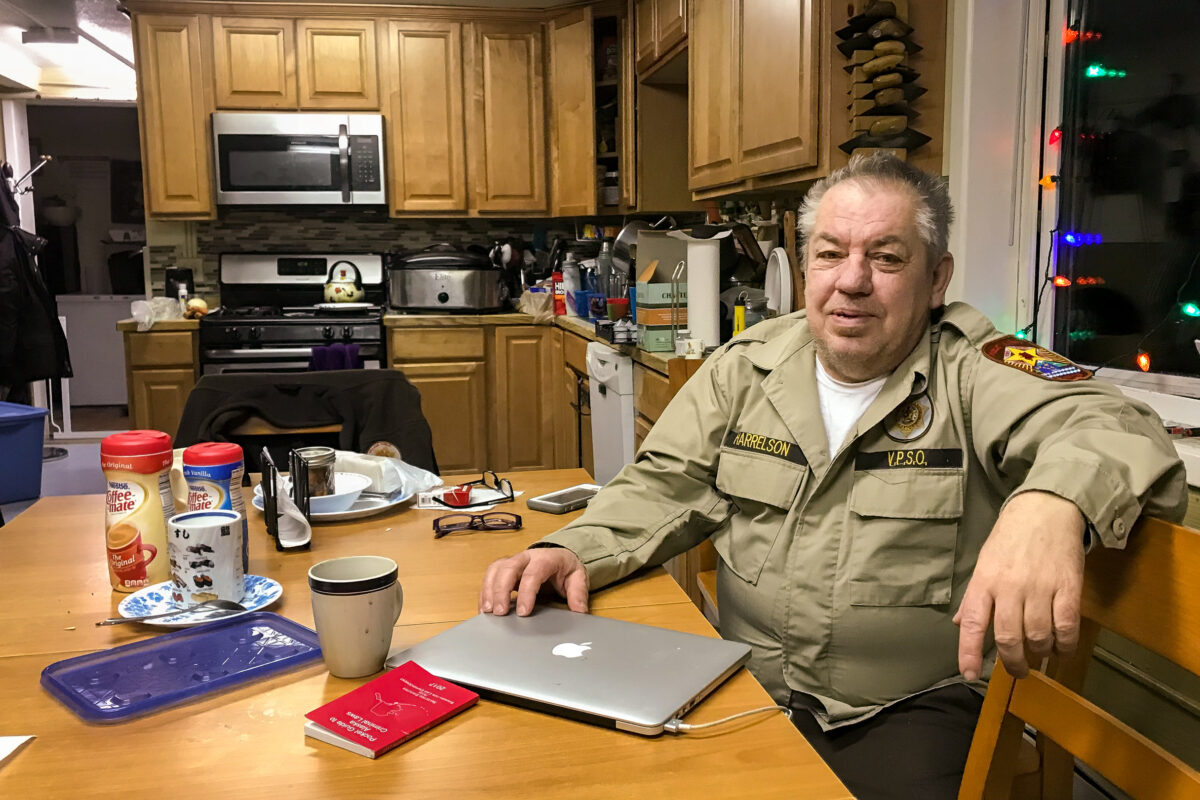 Man in tan security uniform sits at the kitchen table inside his home.