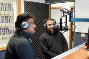 Two priests wearing headphones sit in front of a radio microphone.