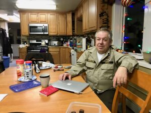 Man in tan security uniform sits at the kitchen table inside his home.