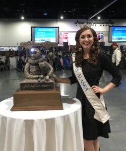Miss Alaska 2017, Angelina Klapperich, poses next to the trophy (bearing a likeness of the late Joe Redington, Sr.) that will be given to the 2018 Iditarod champion.
