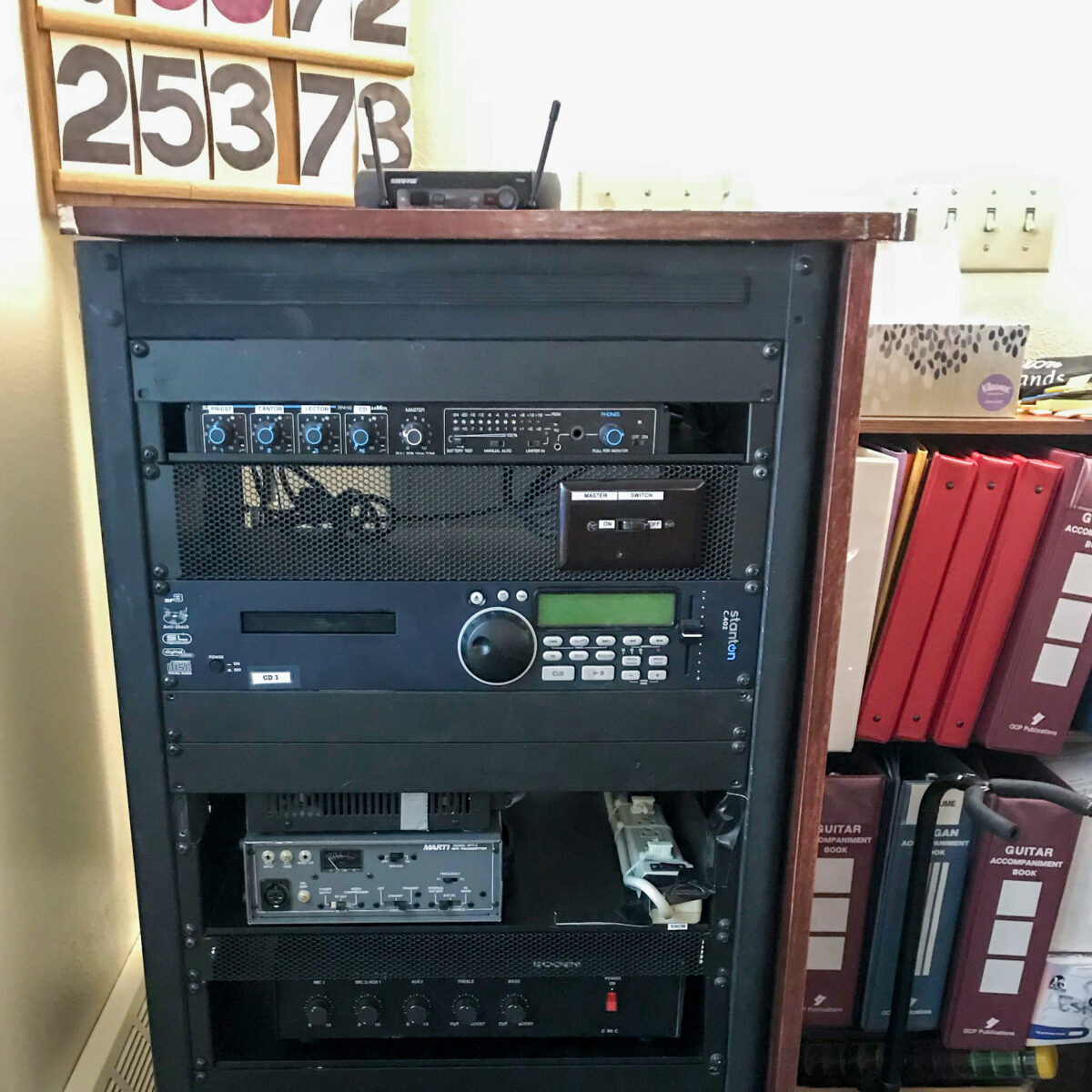In the corner of a Catholic church, a rack of broadcasting hardware, such as a transmitter and CD player.