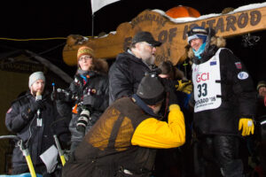 At Iditarod finish line, musher interacts with race officials and smiles for press photos.
