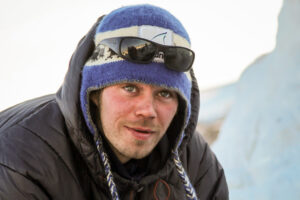 Close-up of musher Joar Leifseth Ulsom, wearing blue hat, sunglasses, black parka