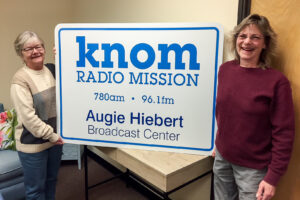 Two women stand next to a large sign reading “KNOM Radio Mission | 780 AM | 96.1 FM | Augie Hiebert Broadcasting Center.”