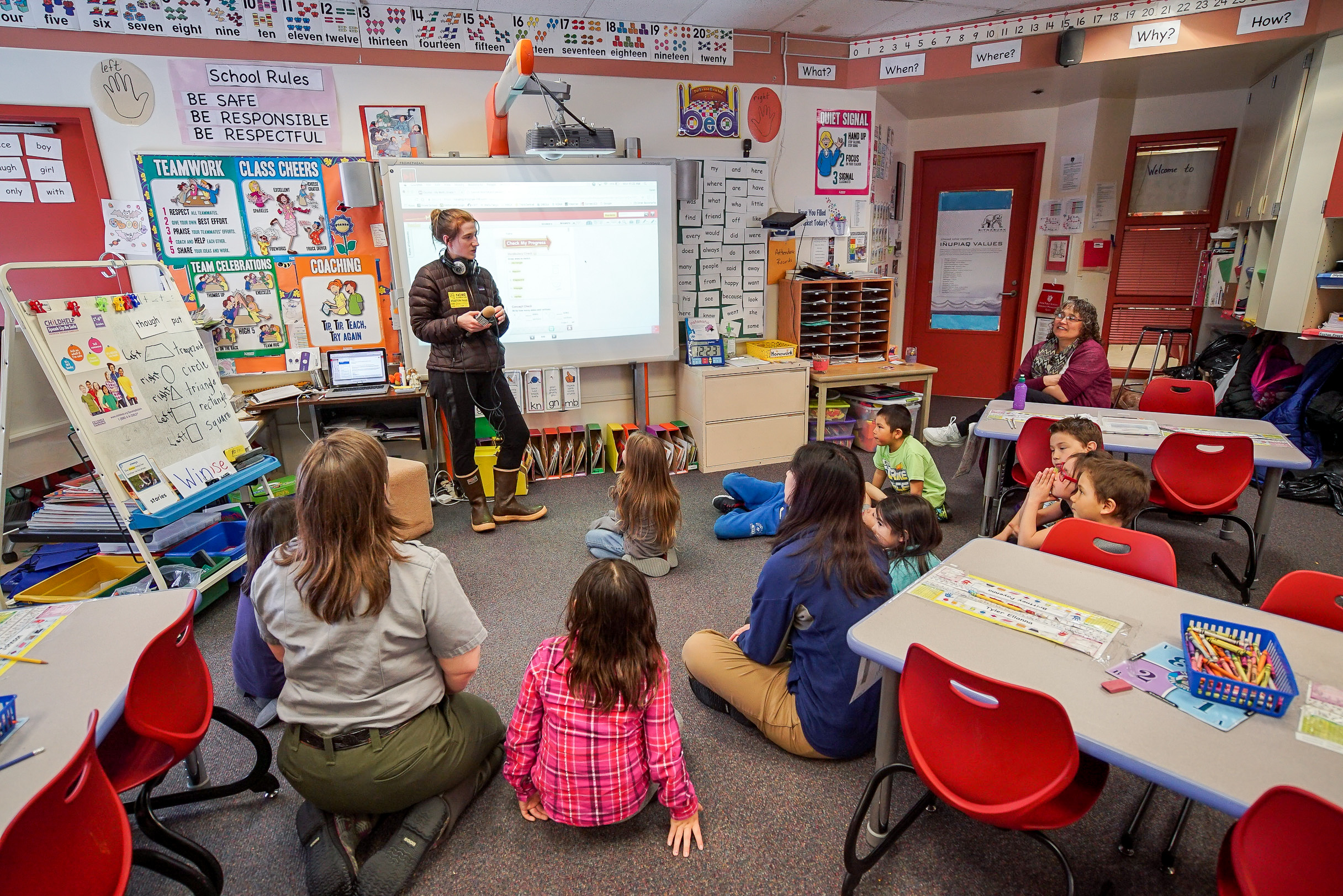 Woman, standing, addresses classroom of elementary school students, their teacher, and a park ranger, all seated.