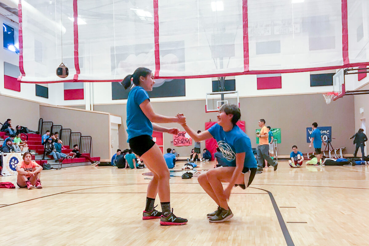 One NYO athlete helps another stand up from the gymnasium floor.