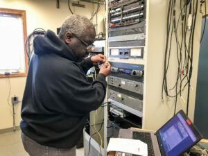 Man pauses to look at laptop while making adjustments to the wiring inside KNOM's AM transmitter building.