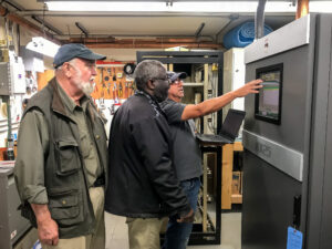 Three men look at a computer screen attached to the newly-installed AM transmitter hardware.