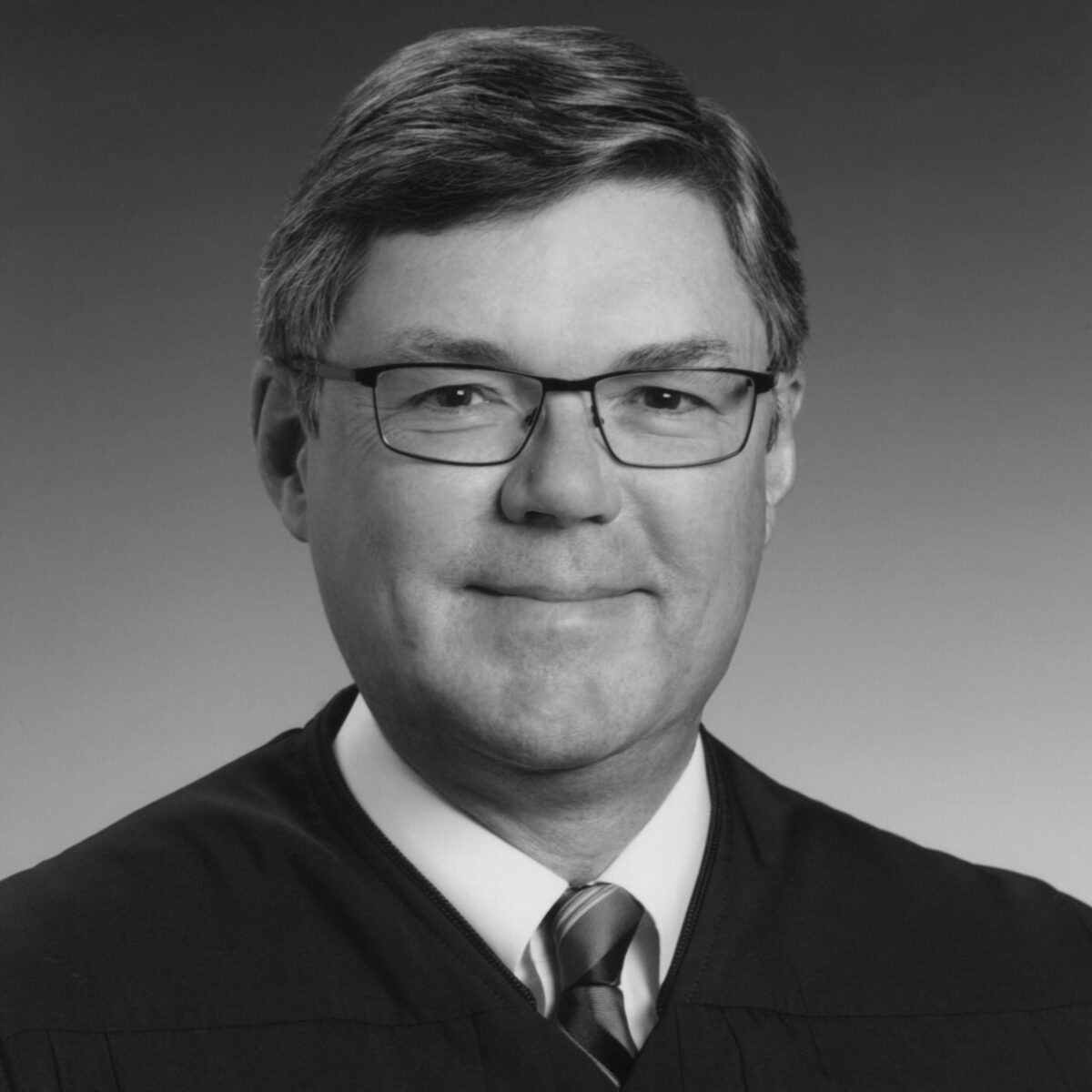 Black-and-white photo of man in glasses wearing shirt and tie and judicial robe.