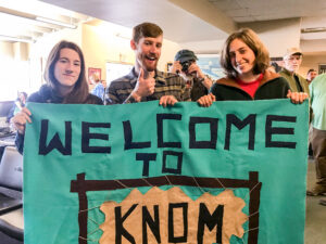 Three people hold a teal "Welcome to KNOM" banner inside a small airport terminal, while a man, standing over their shoulder, holds his own small camera back at the photographer.