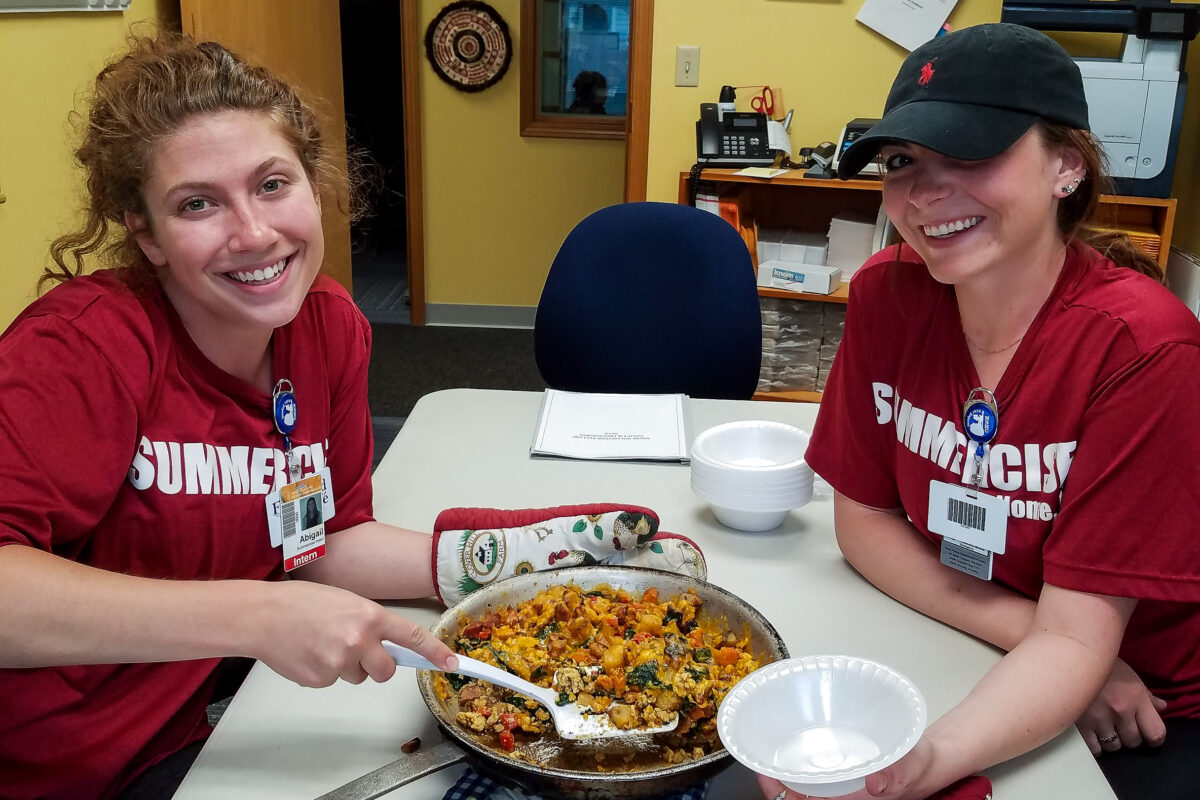Two women in dark red "Summercise" t-shirts smile at camera, sitting at a table inside KNOM studios, while displaying skillet with freshly-cooked egg scramble.