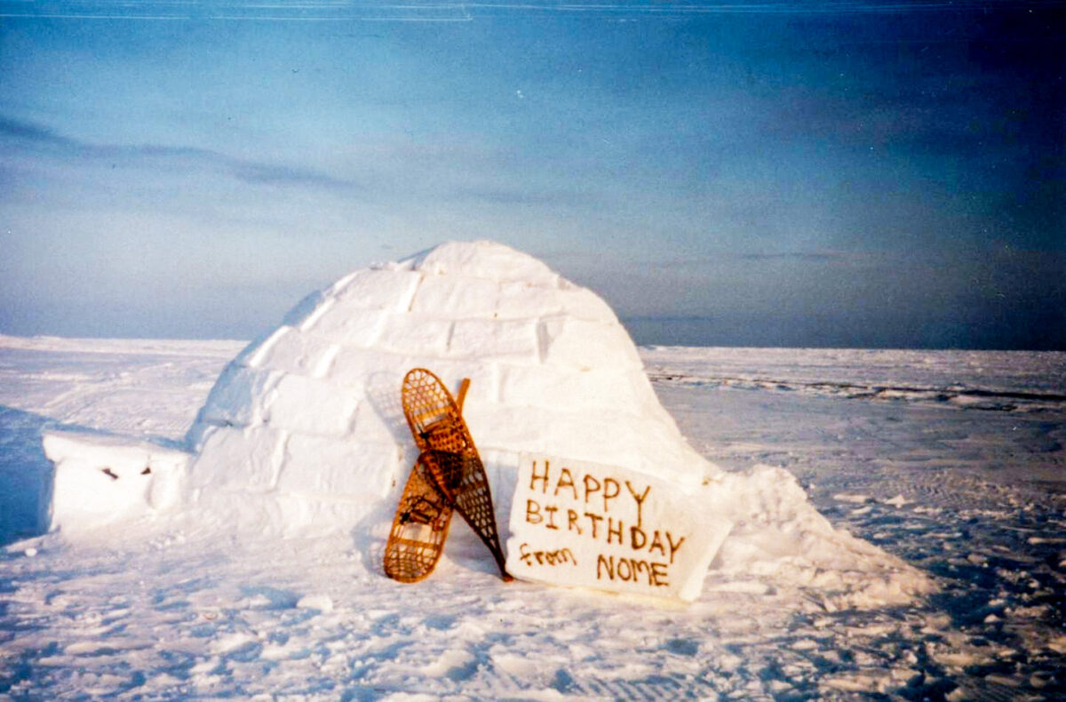 A vintage photo of an igloo on a field of snow and ice, with a pair of snowshoes and a handwritten sign that says “Happy Birthday from Nome.”