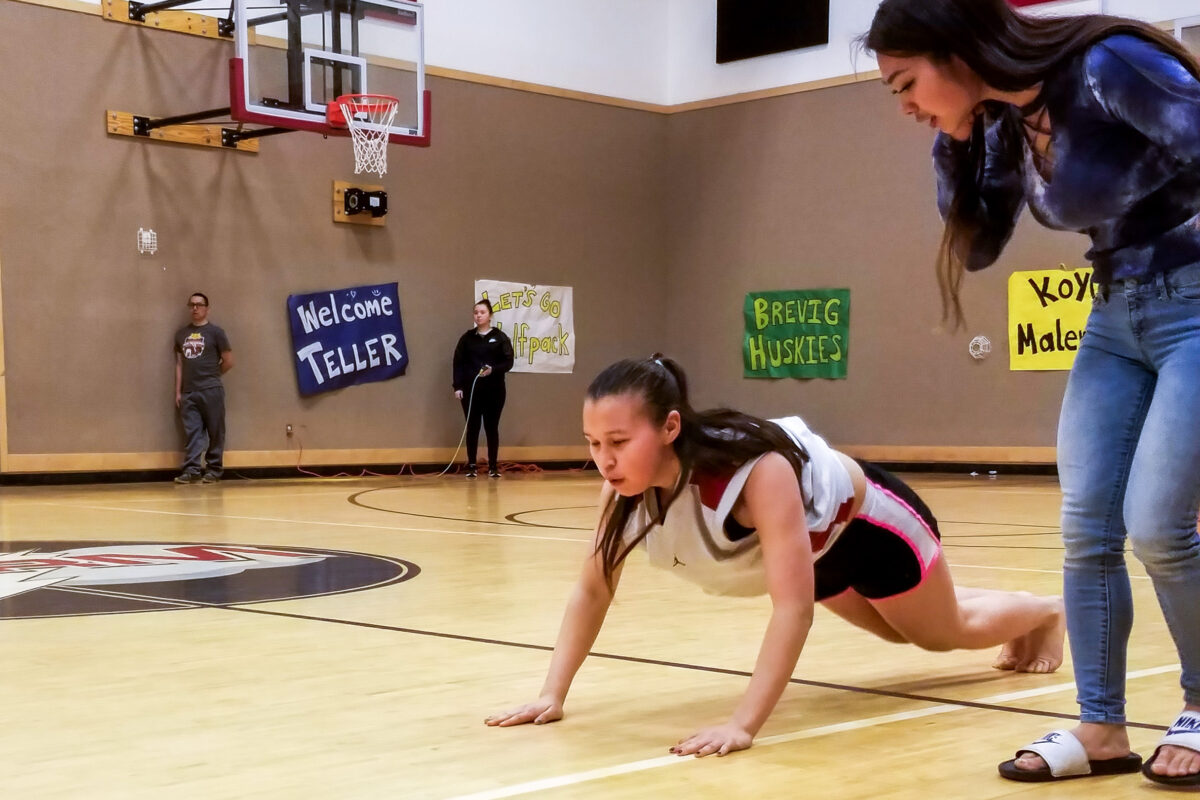 Girl is in position on gymnasium floor similar to a push-up — body almost parallel to the floor, legs behind her, arms extended — while competing in the Native Youth Olympics “seal hop” event.
