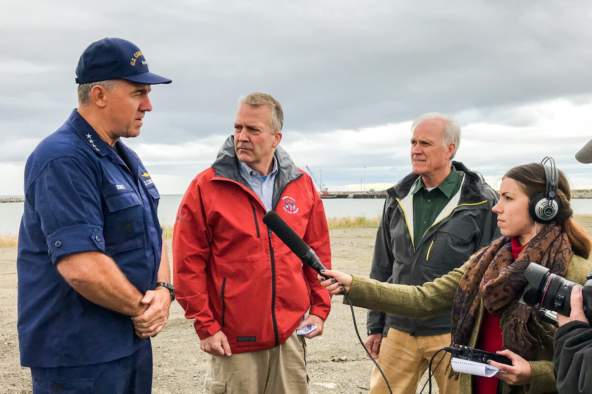 Three men wearing jackets stand on a beach on a cloudy day, talking to reporters.