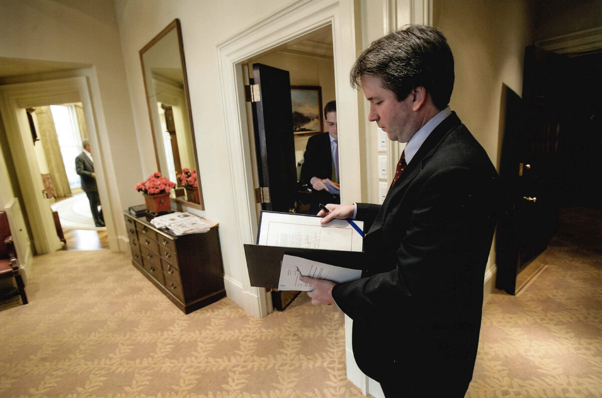 Man in dark suit stands inside the White House, looking at a folder of papers.
