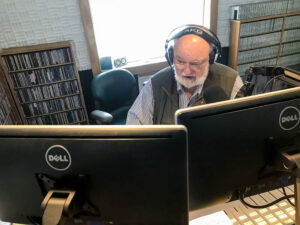 Man with white beard sits at a bank of computer screens inside a radio studio.