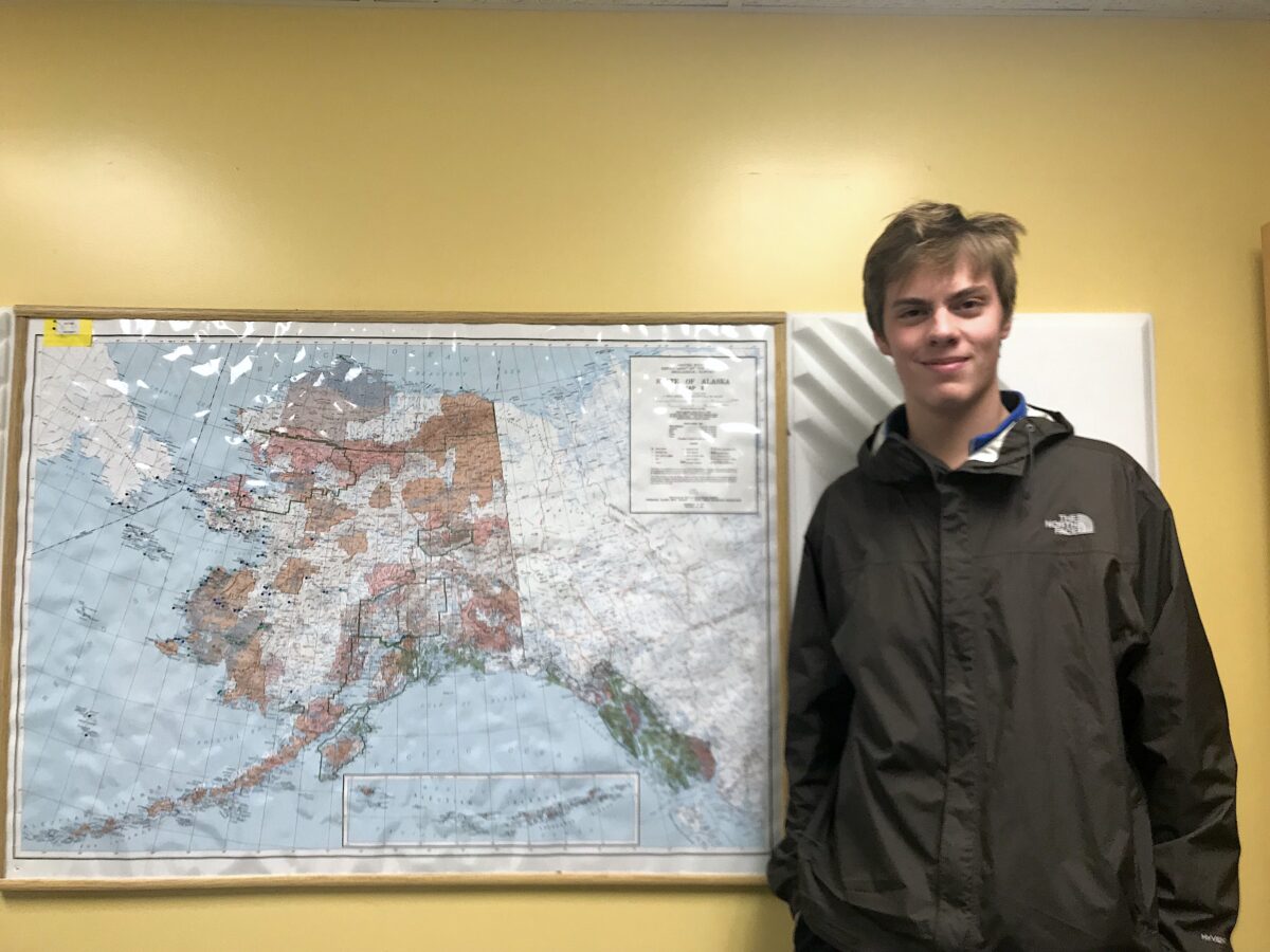 A young man stands next to a map of the state of Alaska in a room with yellow walls.