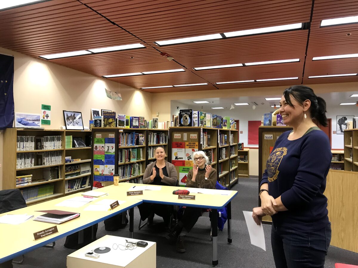 a woman stands smiling looking to the left, as two seated women look on and applaud in a school library.
