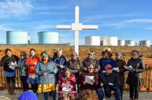 Choir sings in front of large white cross, outside, with large fuel tanks in the background on a sunny autumn day near Nome.