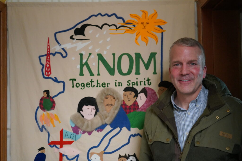 man stands smiling in front of a banner that reads "KNOM, together in spirit" and felt patchwork