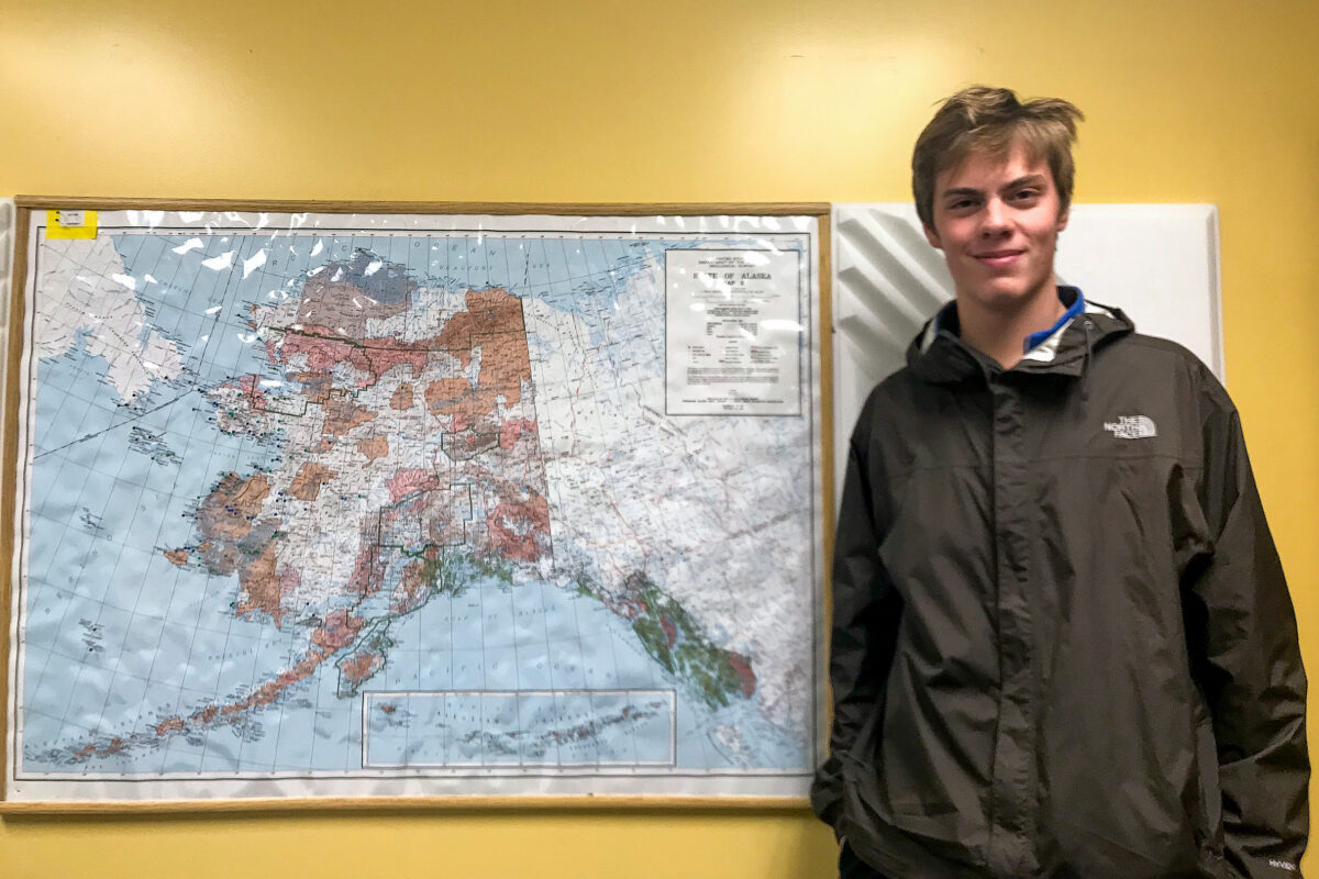 Young man wearing black windbreaker jacket stands smiling next to framed map of Alaska.