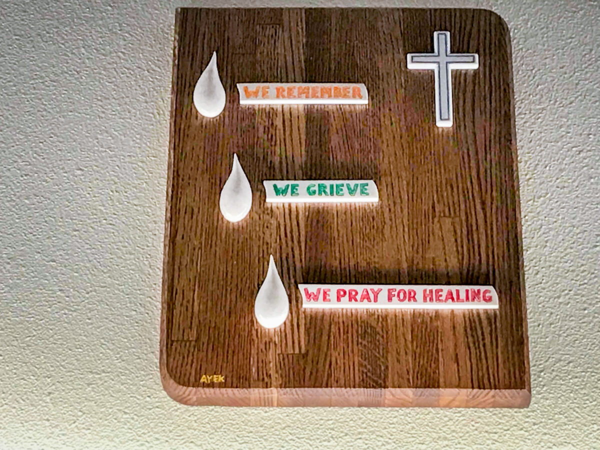 A wooden plaque bearing a simple, white cross, three teardrop shapes, and the words “We Remember, We Grieve, We Pray for Healing.”