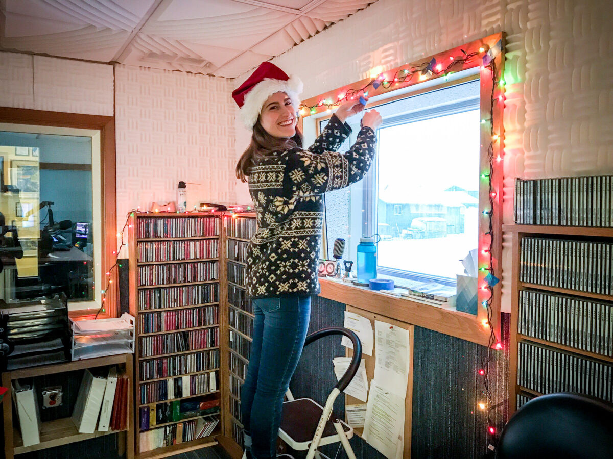 Woman in sweater and Santa hat hangs colored Christmas lights along the window frame of a radio studio.