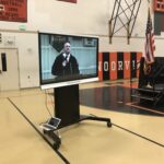 A large screen in a gymnasium depicts a man speaking on a microphone