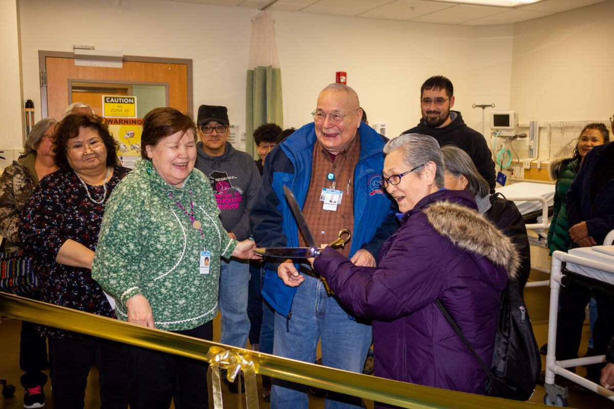 Group of people smiling and laughing inside Nome hospital as woman holds large scissors up to gold-colored ribbon.