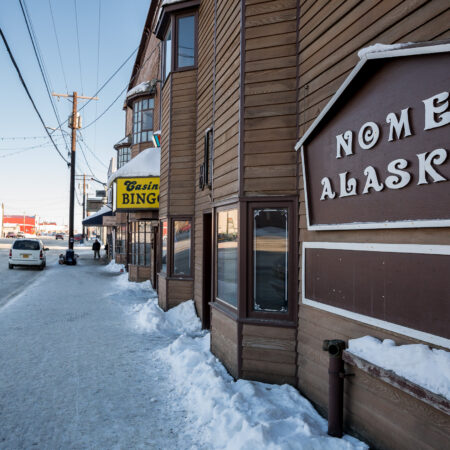 Snowy sidewalk and large sign reading "Nome Alaska" along a wood-paneled building on a winter's day in Nome.