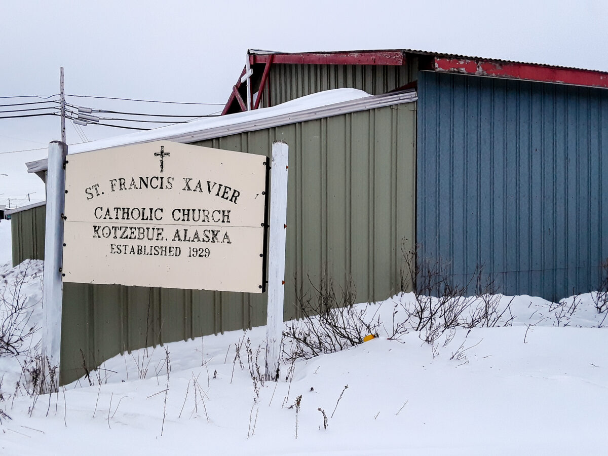 Sign reading "St. Francis Xavier Catholic Church, Kotzebue, Alaska, Established 1929" standing in front of a building with green and blue metal siding. Snow covers the ground.